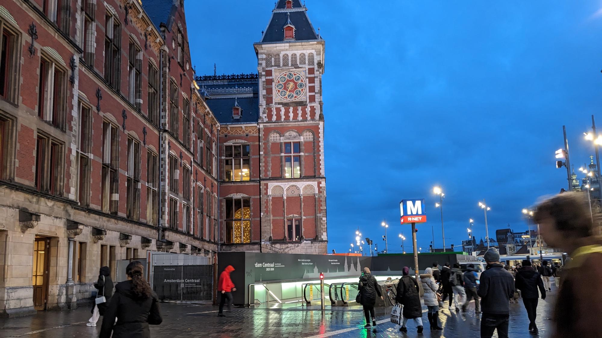 Brick station building with a tower, a pole with an M on a blue cube is in front of the station.